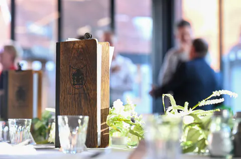 A wooden menu board on the table, guests talking in the background.