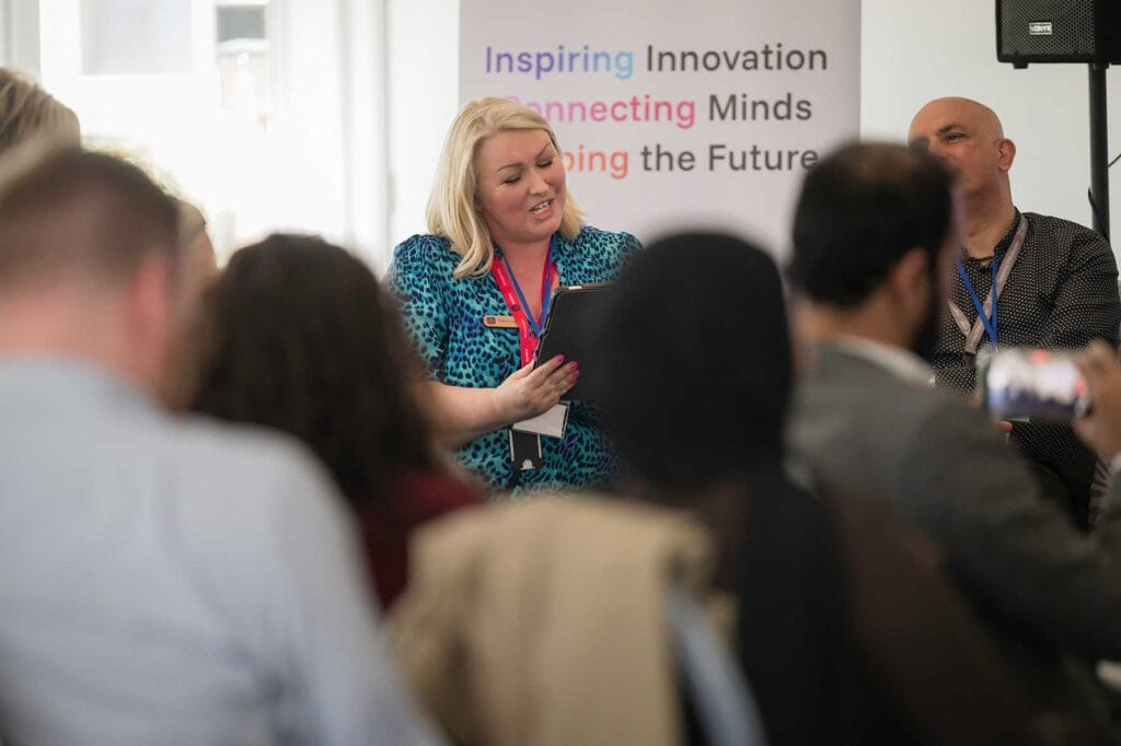 Debbie-Lewis-OVERT-Stem-2 | Business MK Woman presenter in a blue patterned dress holds a tablet while speaking to an audience at a conference, banner reading Inspiring Innovation in the background.
