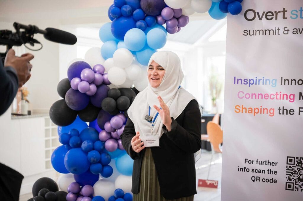Maryam-Jazeem | Business MK Smiling woman in a white hijab speaks at an event beside a blue/purple balloon arch while a camera films her.