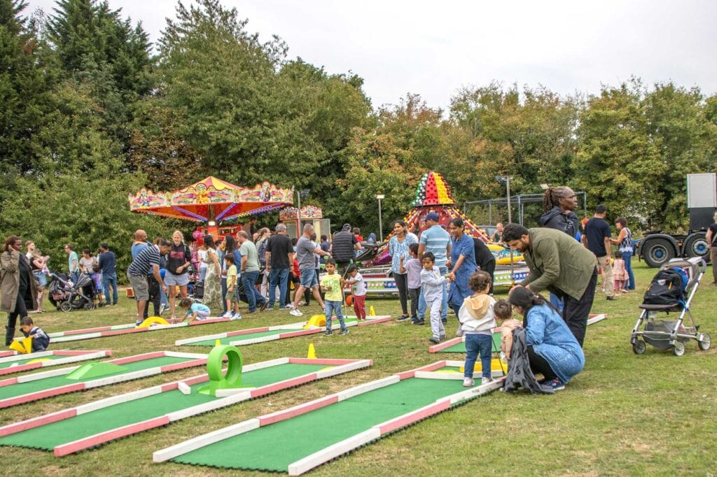 Cranswick-Family-Fun-Day-for-UnityMK | Business MK Crowd of families and children at an outdoor fair with a colorful mini-golf course and amusement rides in the background.