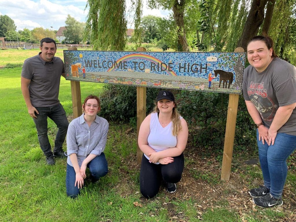 Cranswick-staff-supporting-Ride-High-Groundwork | Business MK Four people posing beside a colorful mosaic sign reading 'Welcome to Ride High' in a grassy park with trees and a rainbow in the background.