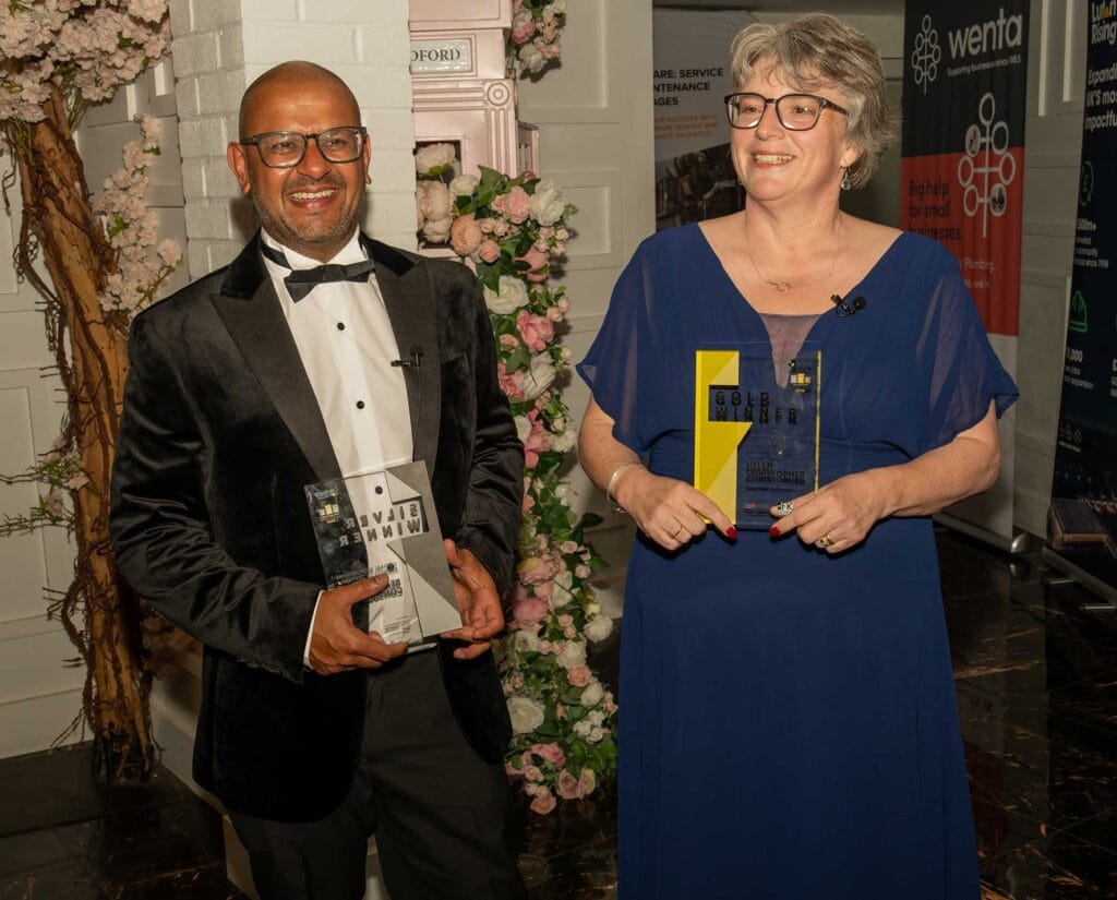 Smiling man in a black tuxedo and glasses holds an award beside a woman in a blue dress who also holds a trophy at an awards event with floral decor behind them.