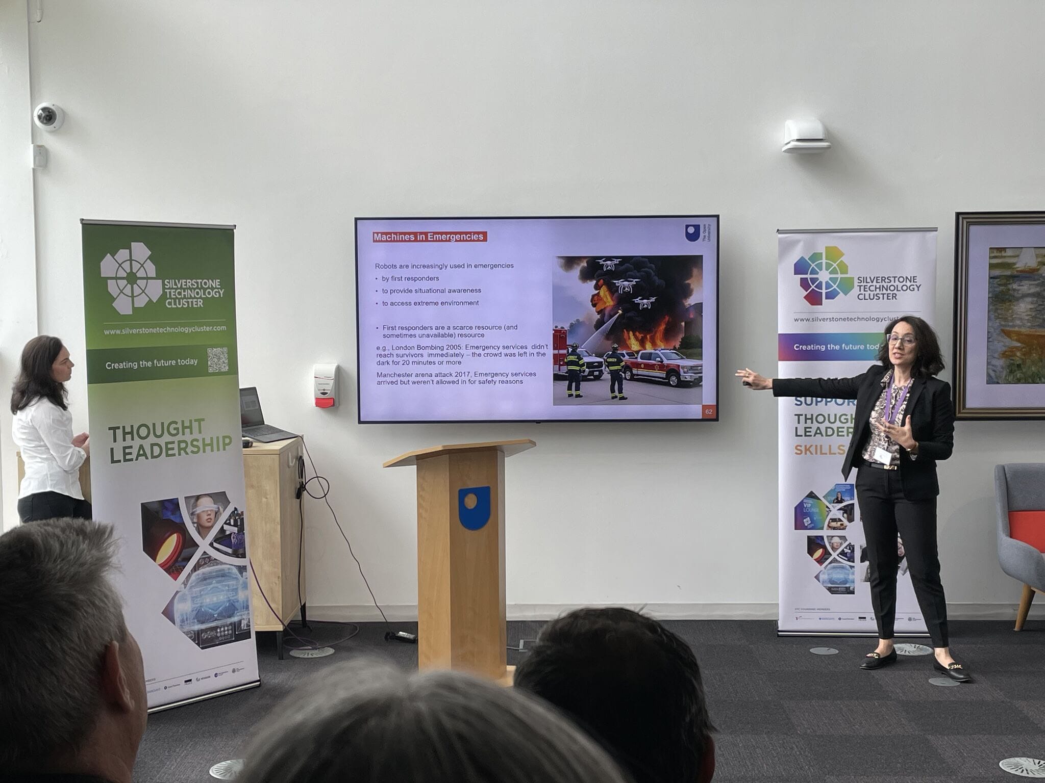 Speaker presenting in a conference room with banners on both sides and a screen showing a slide titled 'Machines in Emergencies'.