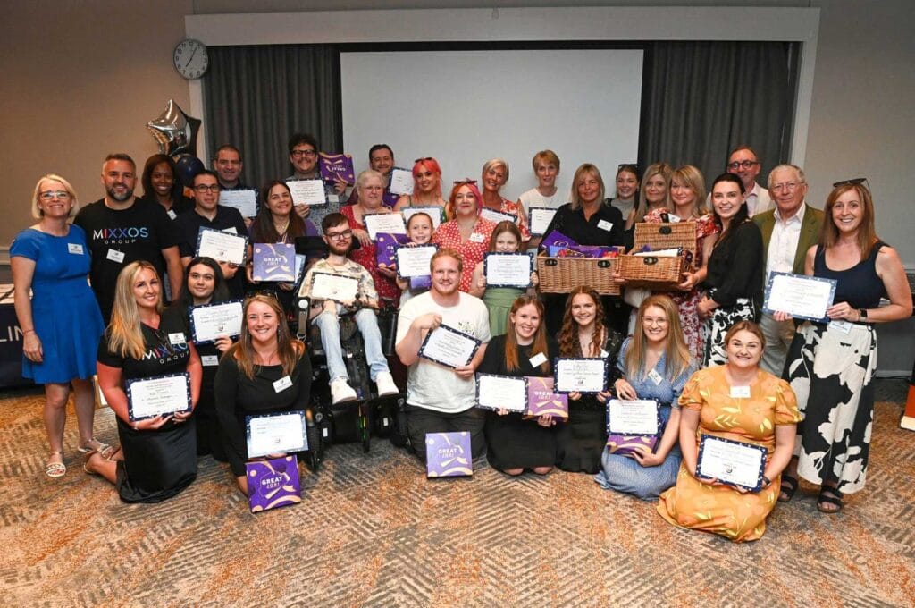 Group-shot-of-winners | Business MK Large group of about 30 people indoors posing for a celebration, many holding certificates and purple gift boxes; balloons and a screen backdrop visible.