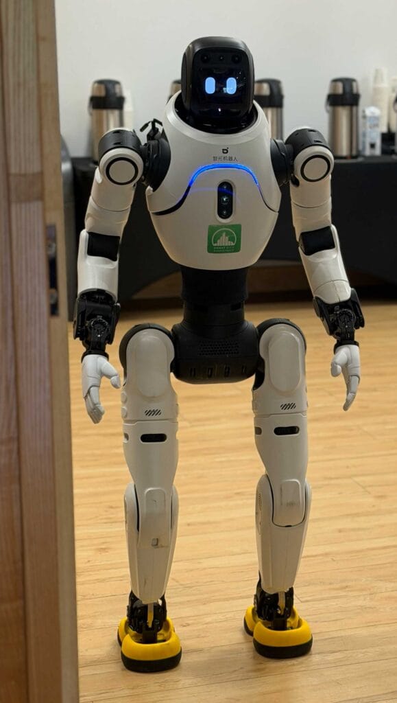 A white humanoid robot standing indoors with blue chest lights, arms at its sides, and yellow-soled feet on a wooden floor.