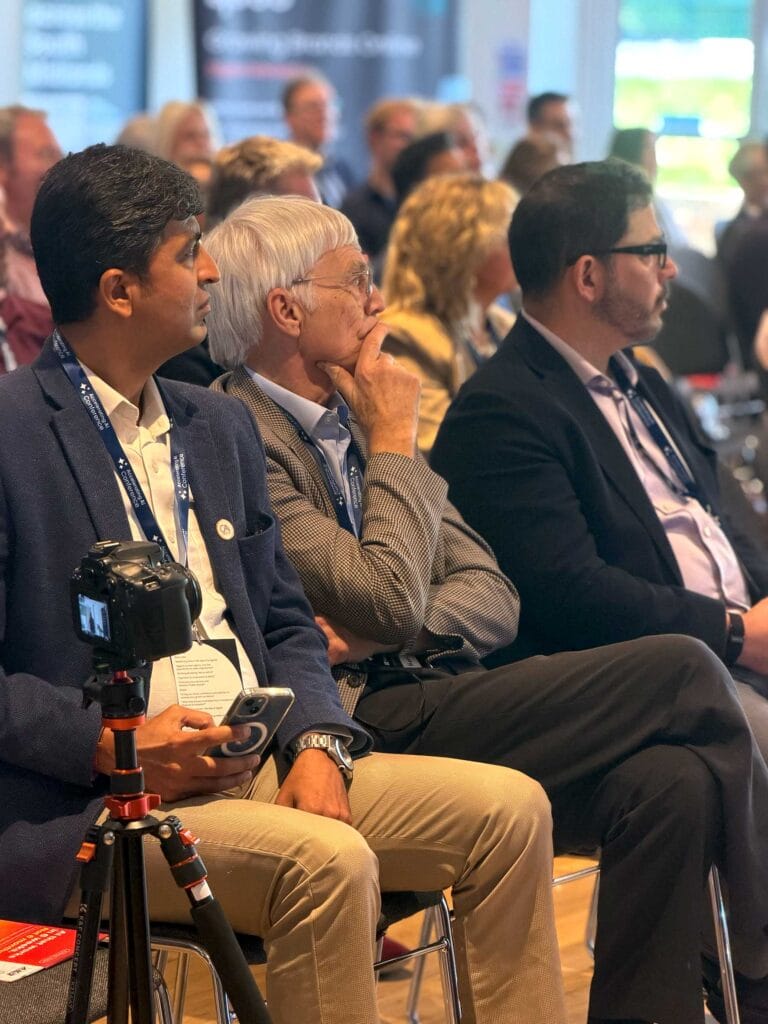Three men in business attire seated at a conference, one with a camera on a tripod, listening to a presentation.