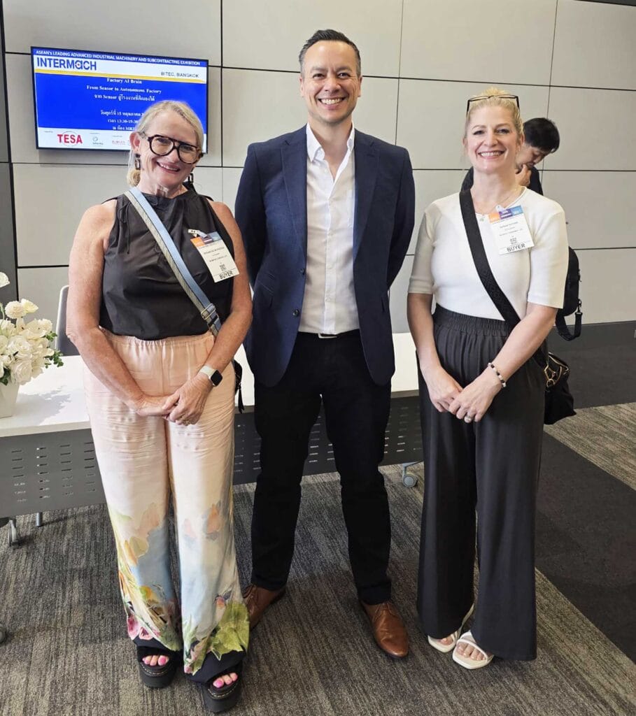 Three people posing at a conference: a woman in floral pants and black top (left), a smiling man in a blue blazer (center), and a woman in a white top with dark wide-leg pants (right), all wearing event badges; background screen visible behind.