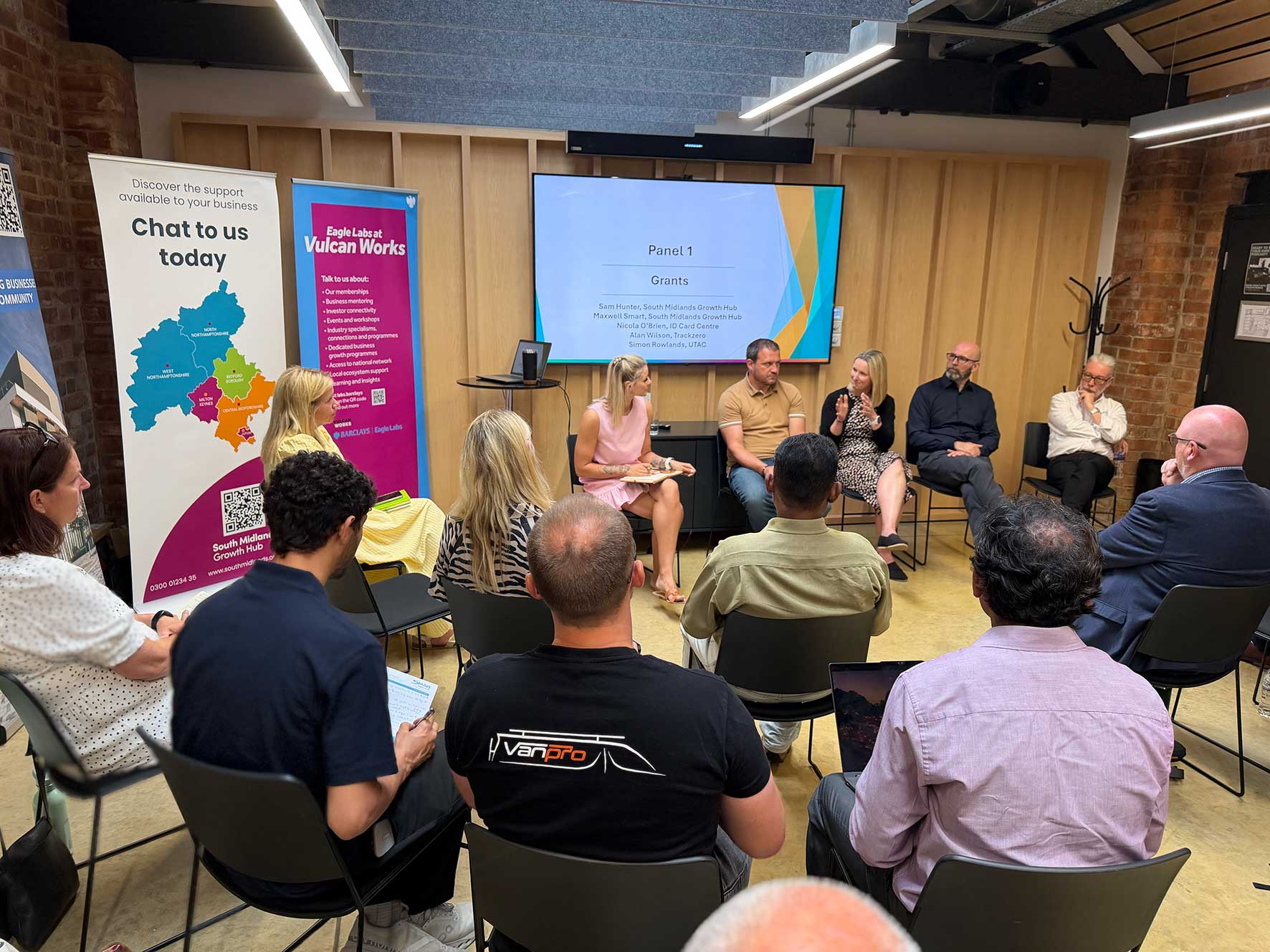 Panel discussion in a conference room with six seated speakers and a large presentation screen reading 'Panel 1 Grants'