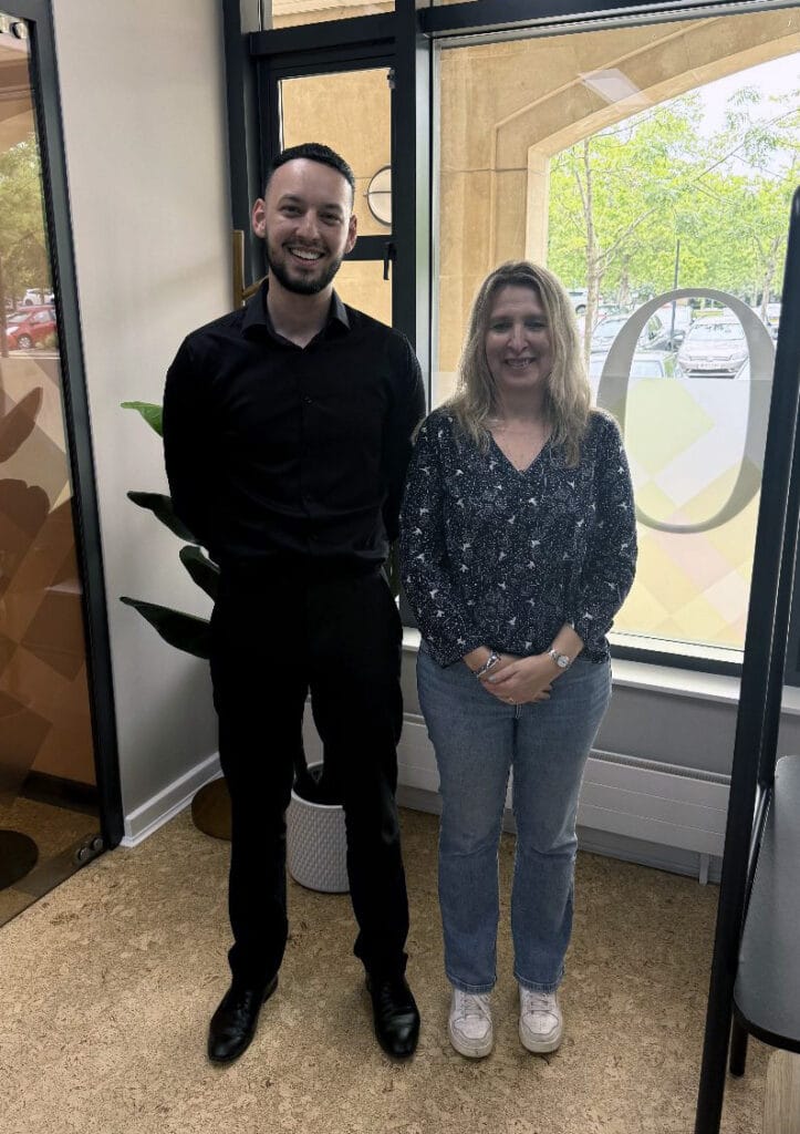 Reece Hussain and Joanna Craig | Business MK Smiling man in black shirt and pants standing next to a woman in a patterned blouse and jeans inside an office. Behind them, large windows and a glass door are visible.