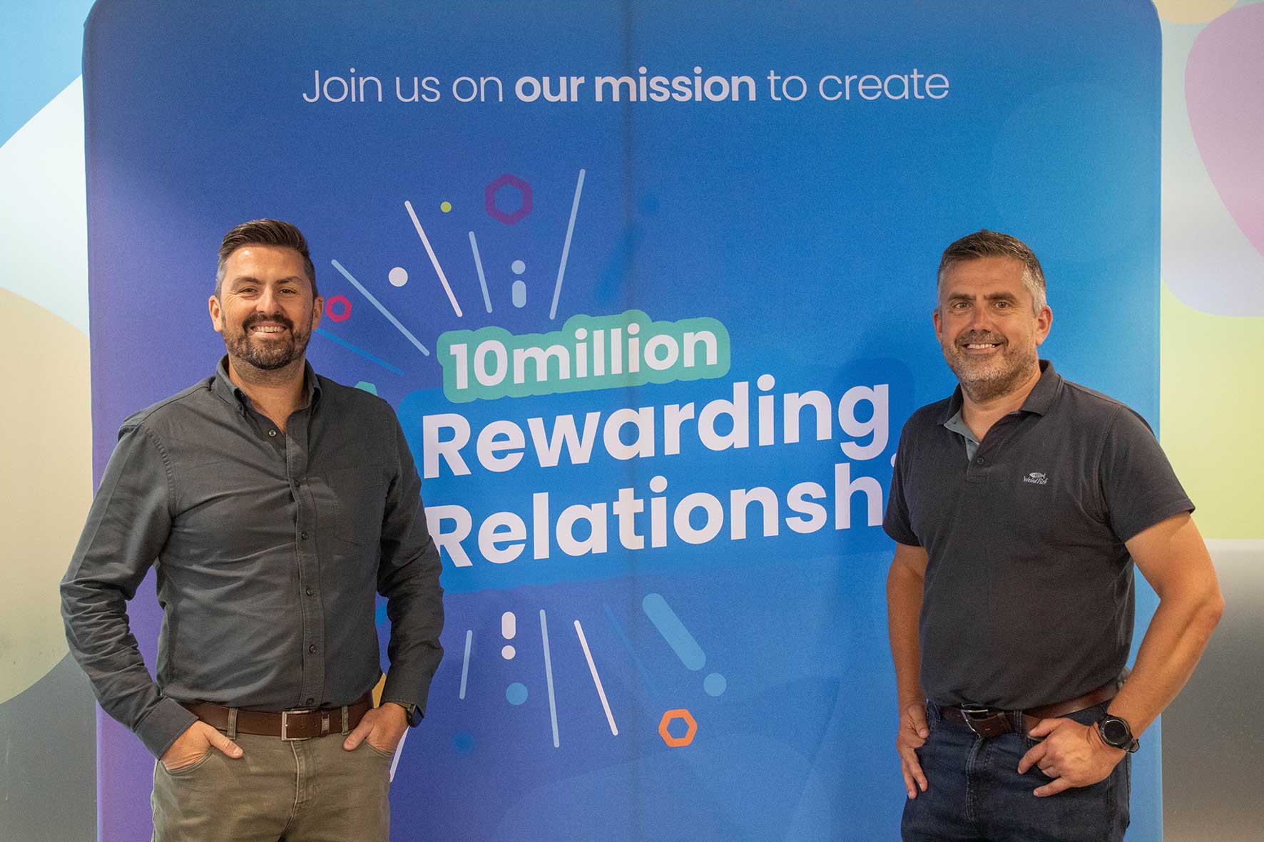 Two men smile and pose in front of a blue banner that says '10 million Rewarding Relationships' and 'Join us on our mission to create'.] ,
