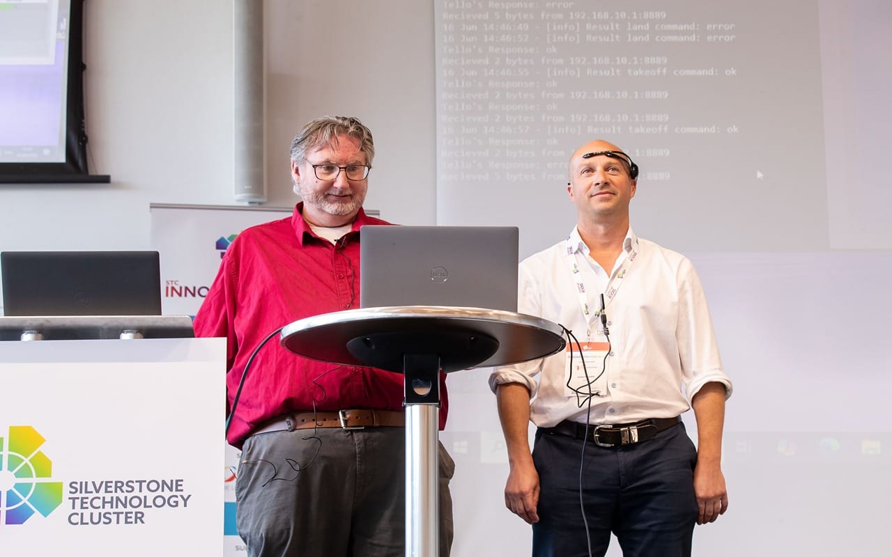 Two men presenting at a tech conference, standing behind a round table with laptops; Silverstone Technology Cluster banner in background.