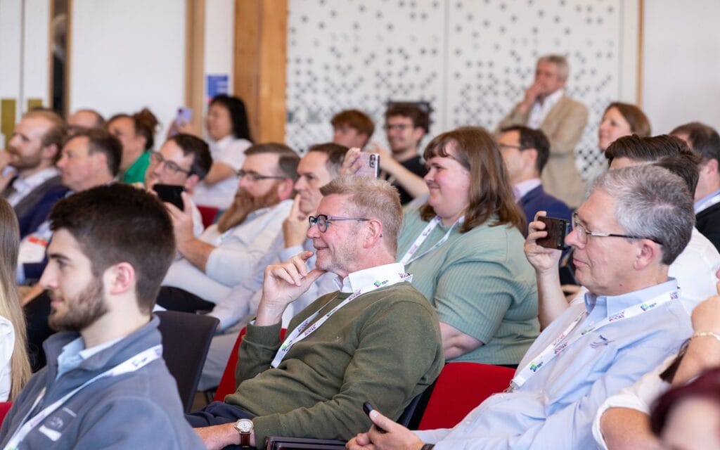Audience of professionals seated at a conference, some taking photos with smartphones.
