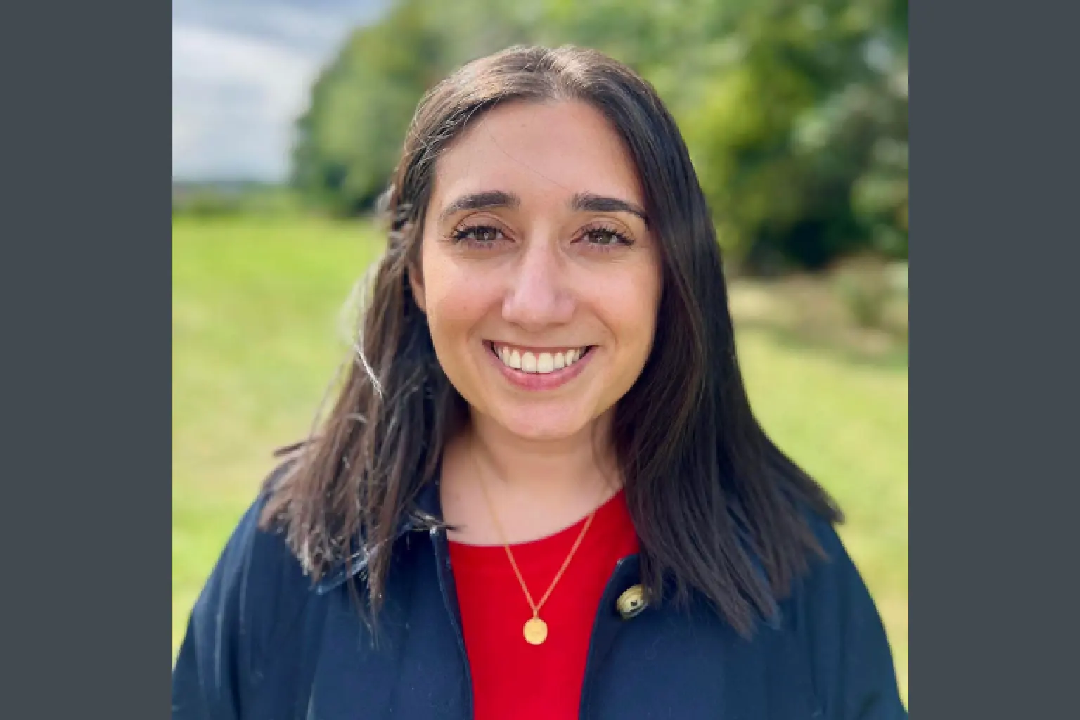 Smiling woman with dark hair wearing a red top and navy jacket outdoors, green field behind her.