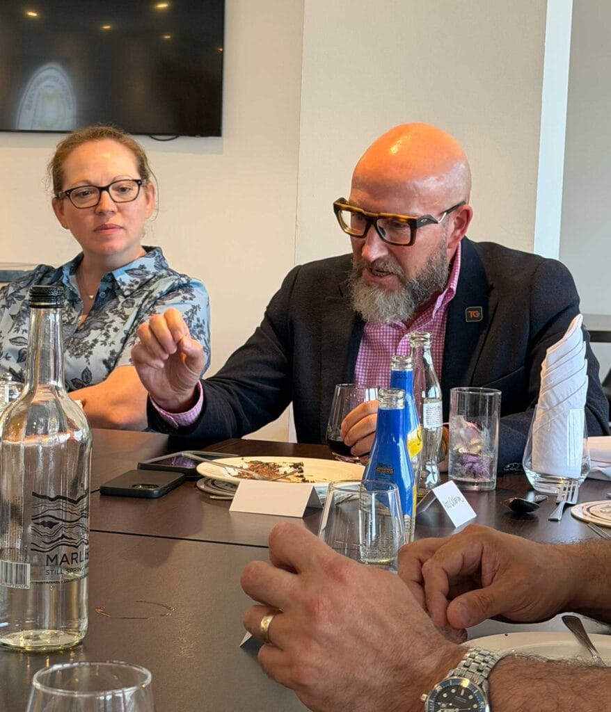 Stephen-Henson | Business MK Two adults sit at a dining table during a meeting, with bottles, glasses, and plates in front of them; a bald man in a blazer and large glasses speaks while a woman with glasses and a floral shirt listens.