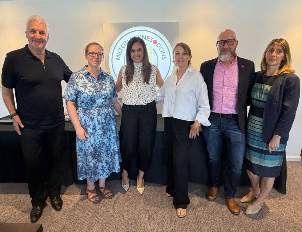 The Partners Behind The Executive Box | Business MK Six adults stand side by side in an office lobby, smiling, with a circular logo on the wall behind them.