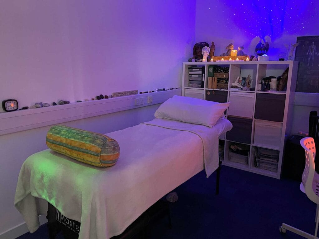 Treatment bed in a purple-lit wellness room with crystals lined along a shelf and a white cubby storage unit topped with candles and decorations.