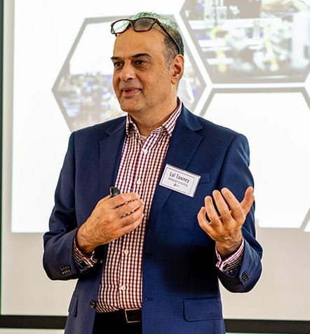 Speaker in a blue blazer presenting, with glasses on his head and a visible name badge, gesturing with his hands.