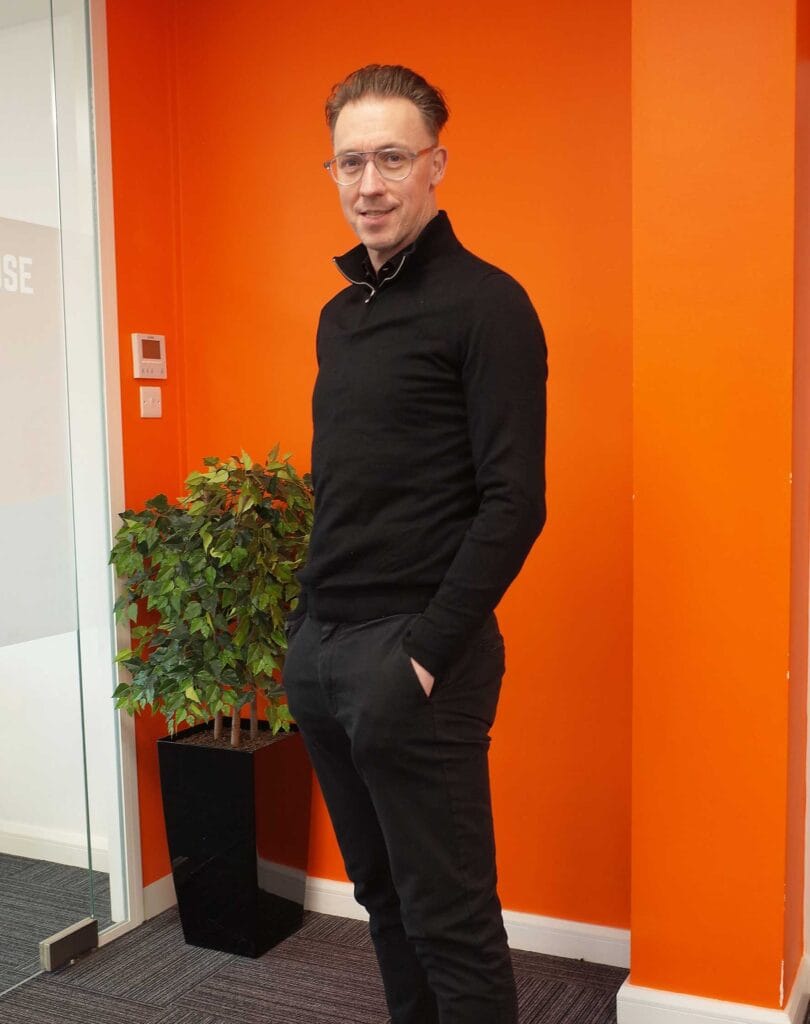 Stephen-Watts-Chief-Operating-Officer-at-TC-Group77 | Business MK Man in a black sweater and glasses stands with hands in pockets against a bright orange wall, near a potted plant in a black planter.