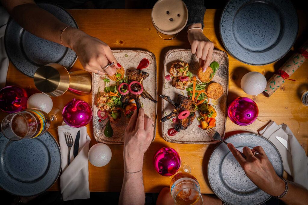 Top-down view of a communal meal with two trays of colorful food and hands cutting and plating at a wooden table.