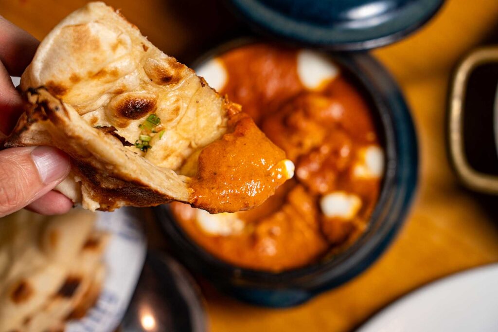 Person tearing off naan bread and dipping it into a creamy orange curry with white dollops and green herbs in a pot.