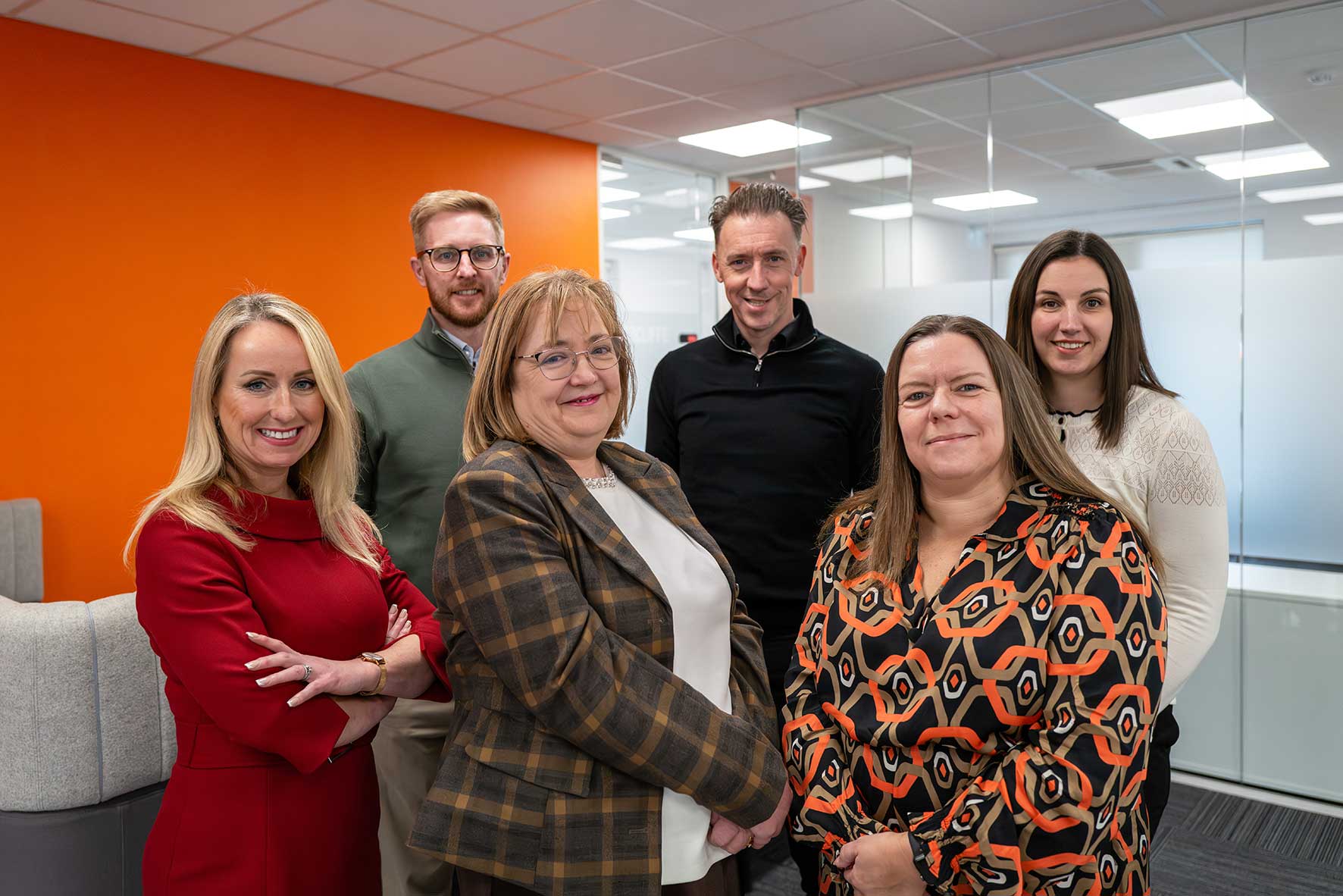 Group of six colleagues posing for a photo in a modern office, orange wall behind them and glass partitions in background, smiling at the camera.