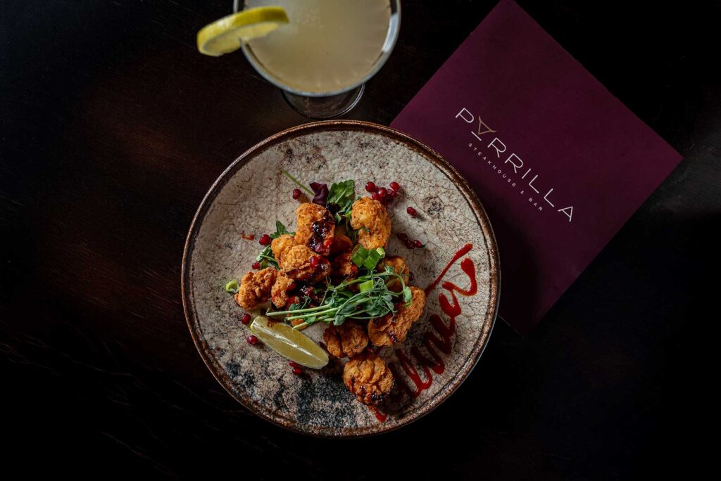 parrilla-christmas-dining-milton-keynes | Business MK Plate of crispy fried chicken bites with greens, pomegranate seeds, and a lemon wedge on a rustic plate; dark table and a burgundy menu nearby saying PYRILLA.