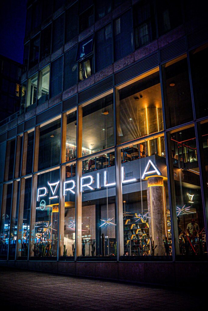 parrilla-the-hub-milton-keynes-restaurant | Business MK Night view of a glass-fronted building with illuminated interiors and neon signage visible through tall windows.