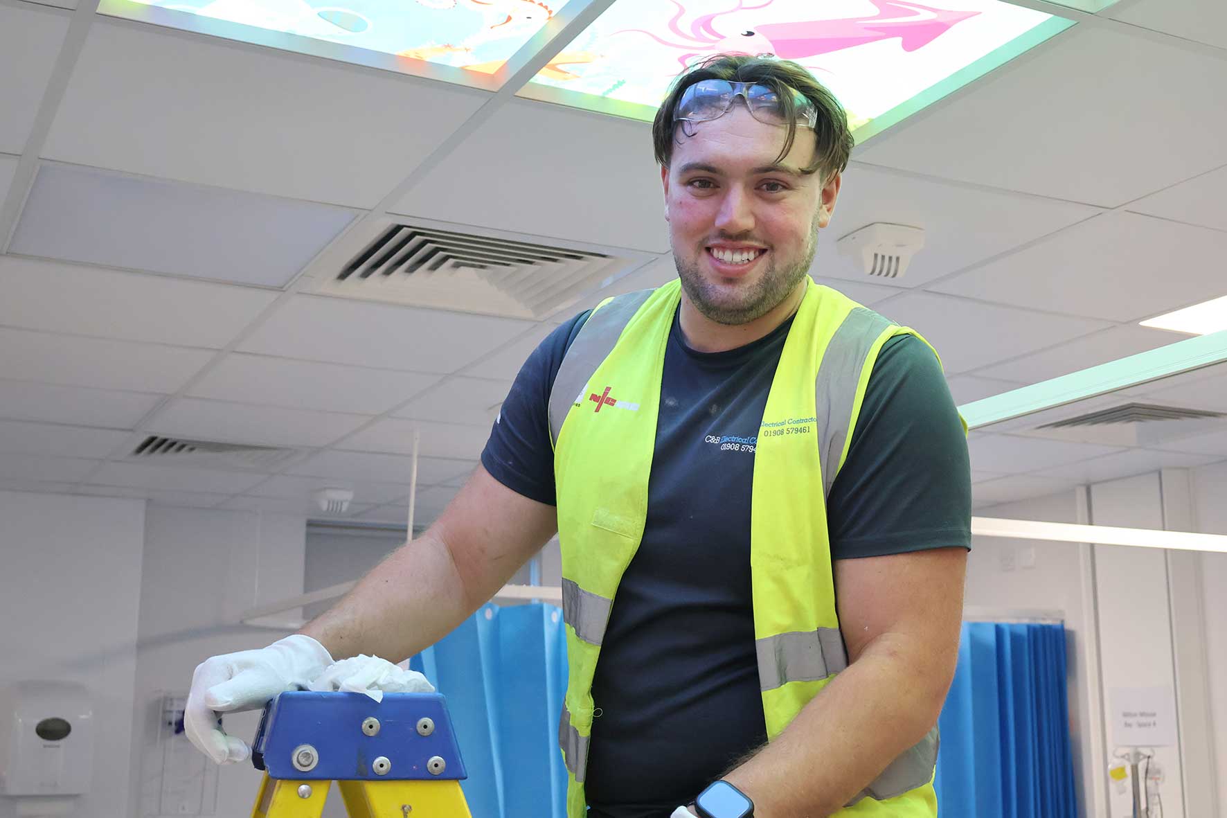 Man wearing C&B Electrical Contractors workwear beside a stepladder beneath illuminated ceiling panels.