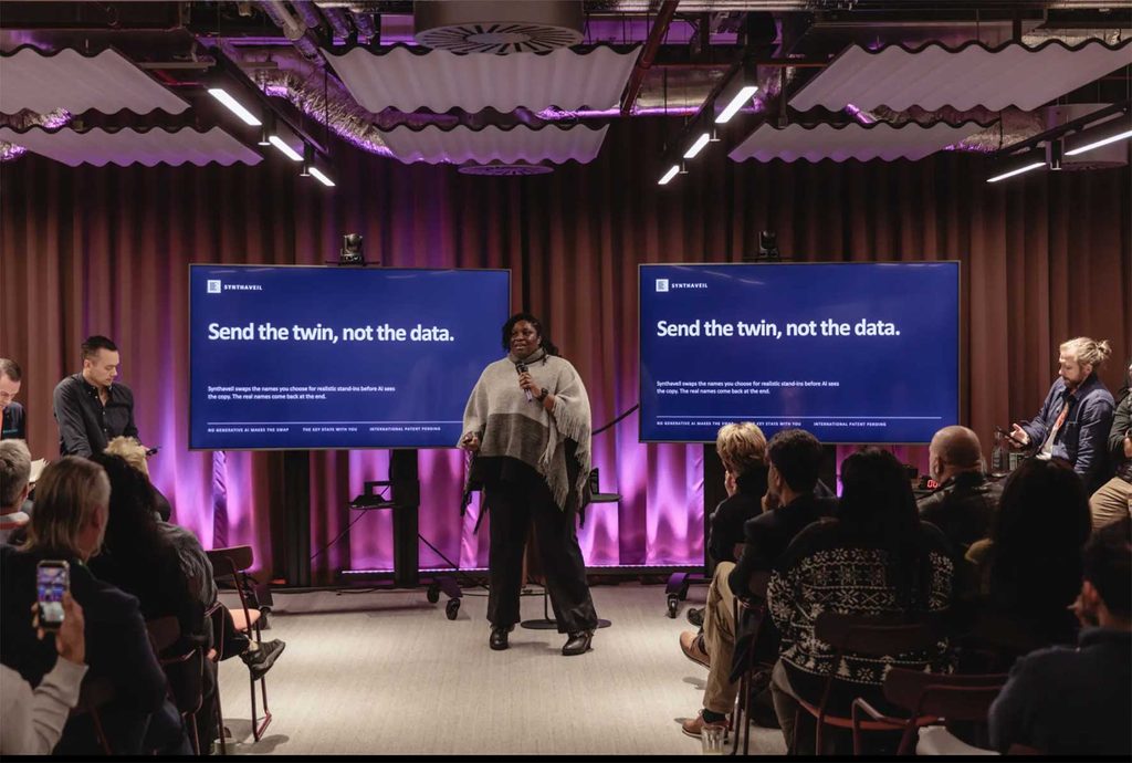 Speaker with microphone on a stage between two large blue presentation screens that read, 'Send the twin, not the data.' Audience seated in front in a dimly lit room with purple uplighting.