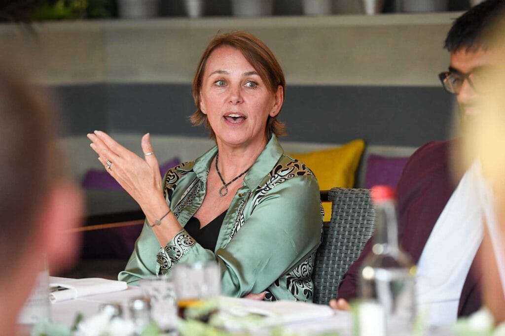 Woman with short brown hair speaks during a meeting, gesturing with her left hand at the table.