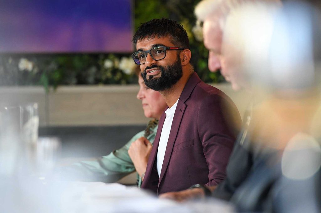 Man with glasses and beard in a burgundy blazer speaking during a meeting; teammates are blurred in the background.