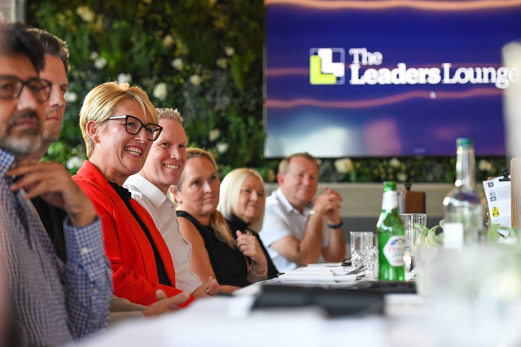Group of professionals seated along a long table, smiling and engaging in a discussion, with a 'The Leaders Lounge' sign in the background.