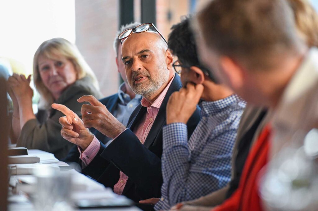 Group of professionals at a long table in a meeting, the man in the center gestures with hands while others listen.